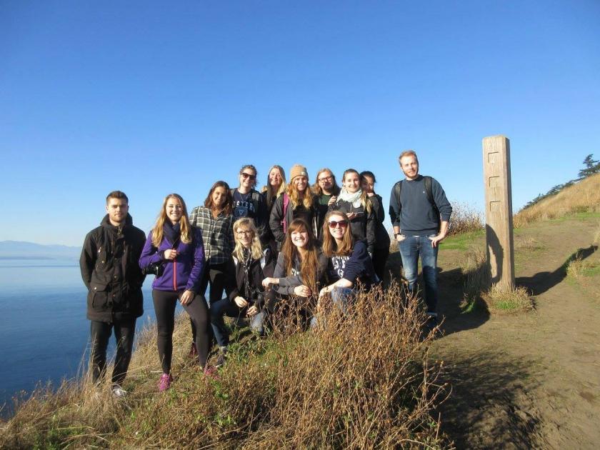 International Buddies (group of 13 students) at Ebey's Landing standing in the sunshine off the side of a trail smiling at the camera