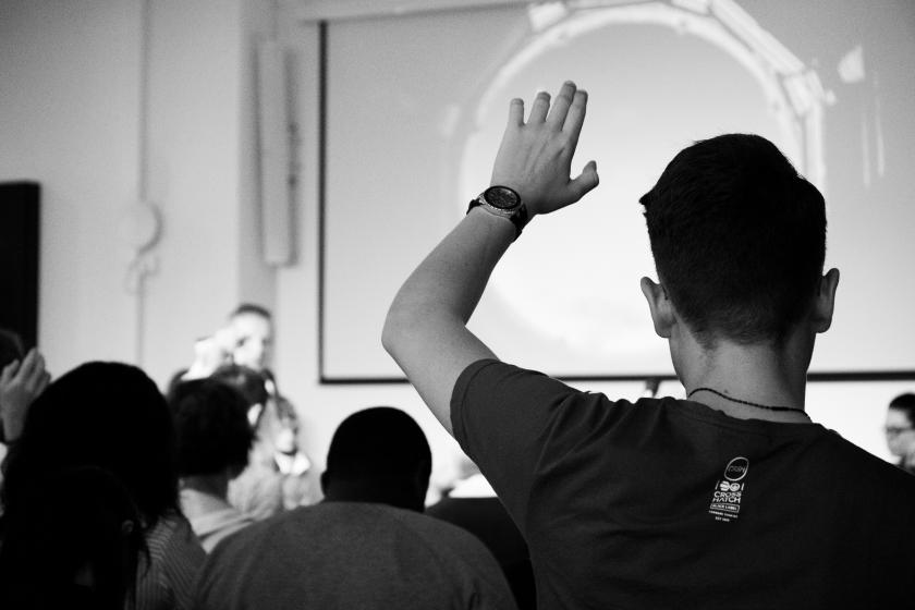 black and white photo of the back of a student raising their hand. Photo by Felicia Buitenwerf, Unsplash