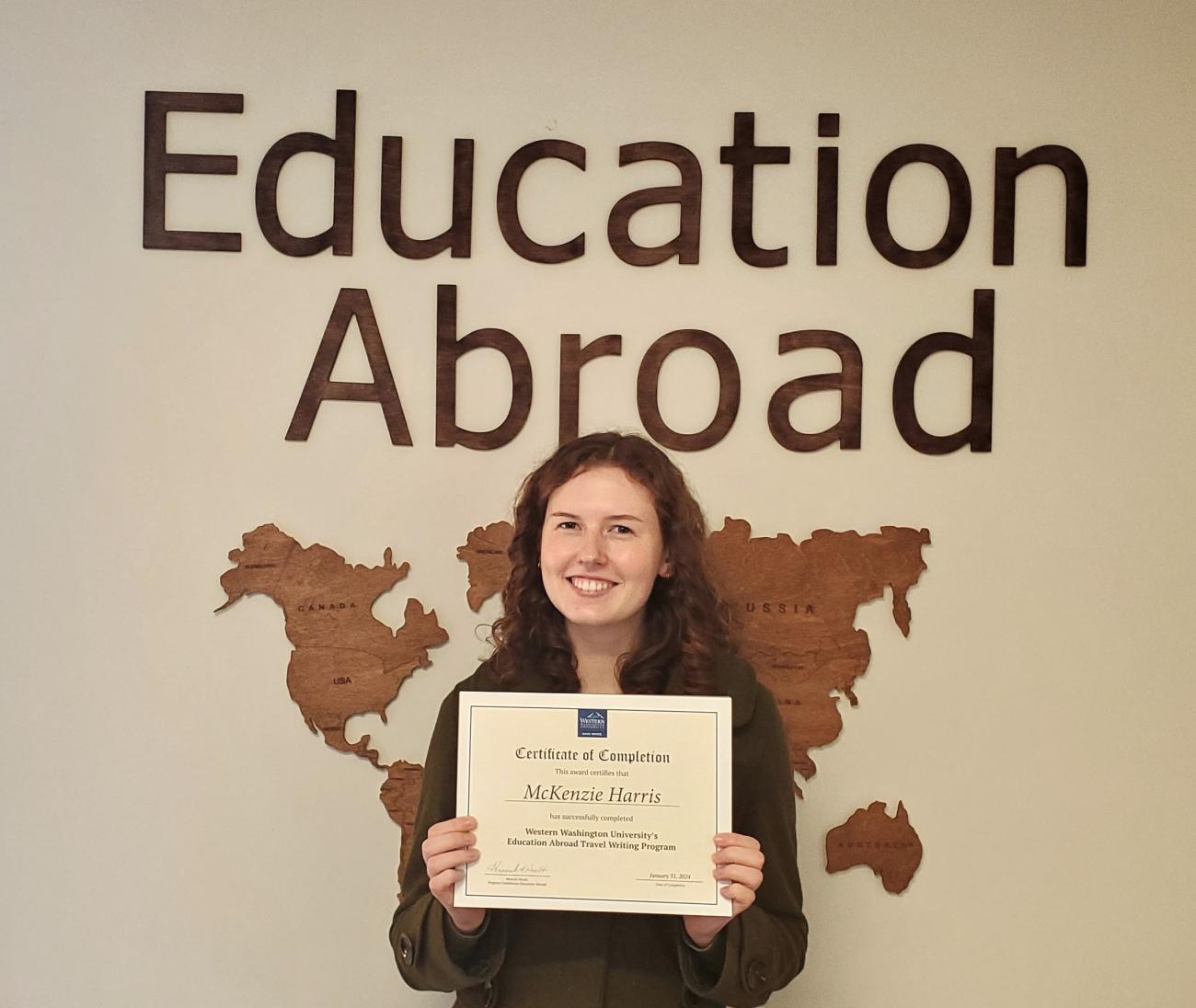 student standing in front of Education Abroad office sign, holding Travel Writing Certificate